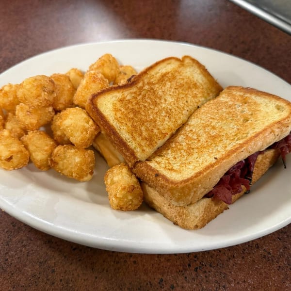 A grilled pastrami sandwich with golden tater tots on a white plate at Dempsey's Breakfast and Lunch.